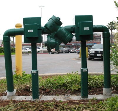 A green metal irrigation control valve assembly with pipes, gauges, and control boxes installed on a concrete pad next to bare ground.
