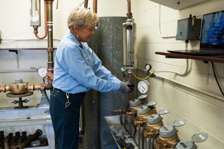 Person in a blue uniform operates gauges and controls in an industrial utility room with pipes, meters, and a wall-mounted monitor.