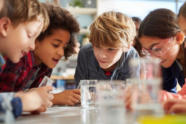 A group of children gather around a table conducting a science experiment with water in a classroom setting.