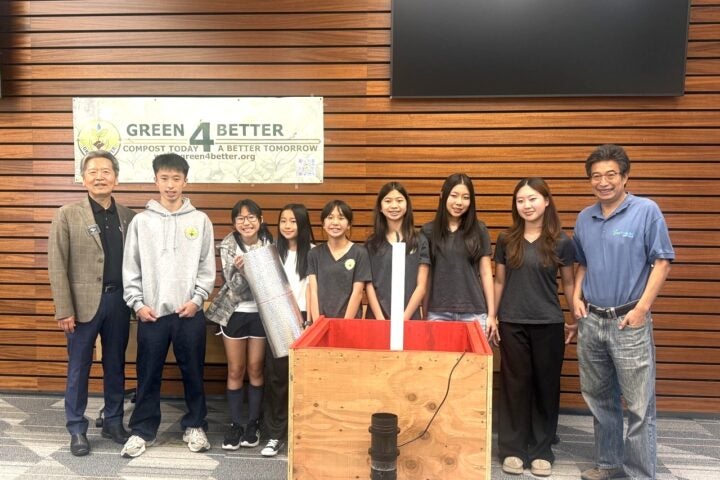 Eight people stand indoors near a composting display bin and a 