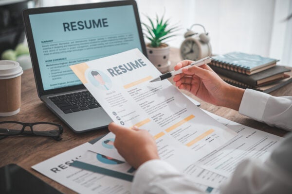 Person reviewing printed resumes at a desk with a laptop displaying a resume, a coffee cup, plant, and clock in the background.