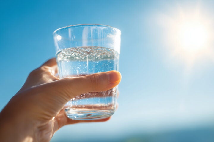 A hand holds a clear glass of water against a bright blue sky with the sun shining in the background.