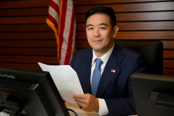 A man in a suit sits at a desk holding papers, with an American flag and computer monitors in the background.