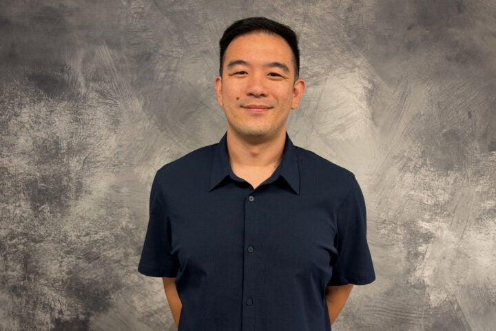 A man with short dark hair stands in front of a gray textured background, wearing a navy blue collared shirt and smiling with his hands behind his back.