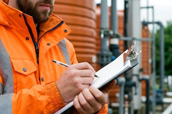 Person in an orange safety jacket writes on a clipboard at an industrial site with pipes and storage tanks in the background.