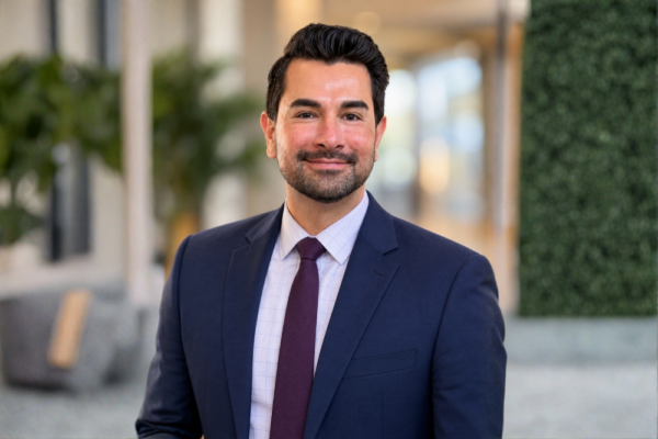A man with a short beard wearing a navy blue suit, light-colored patterned shirt, and a dark tie.